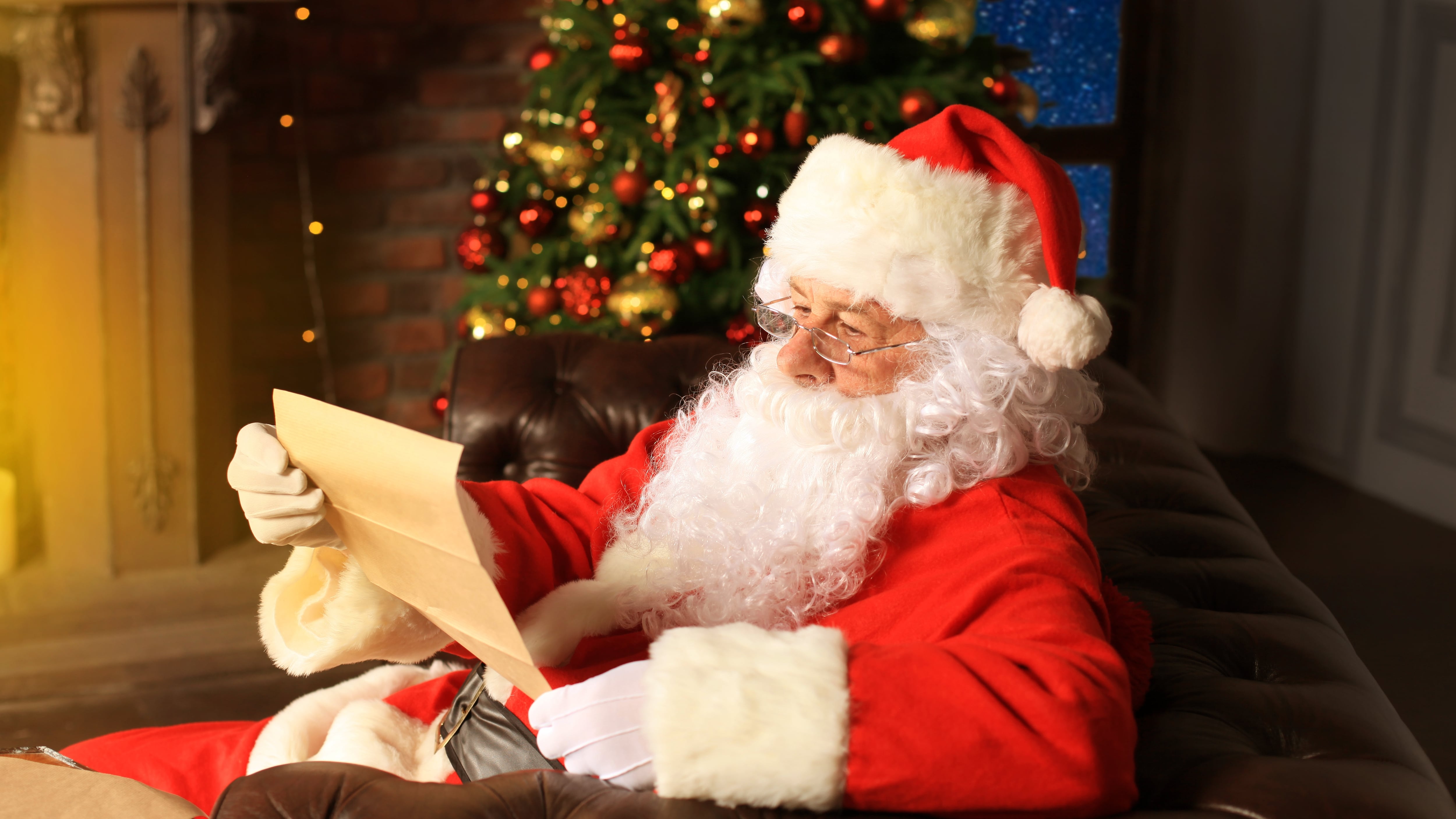 Portrait of happy Santa Claus sitting at his room at home near Christmas tree and reading Christmas letter or wish list